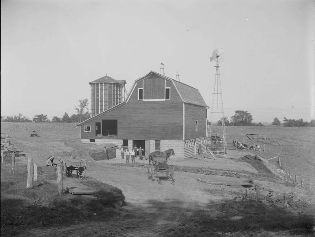 Some people and horses standing in front of a barn, with cows on the hillside next to the barn.